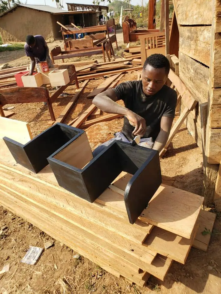 A carpenter painting a pair of wooden shelves outside a timber workshop